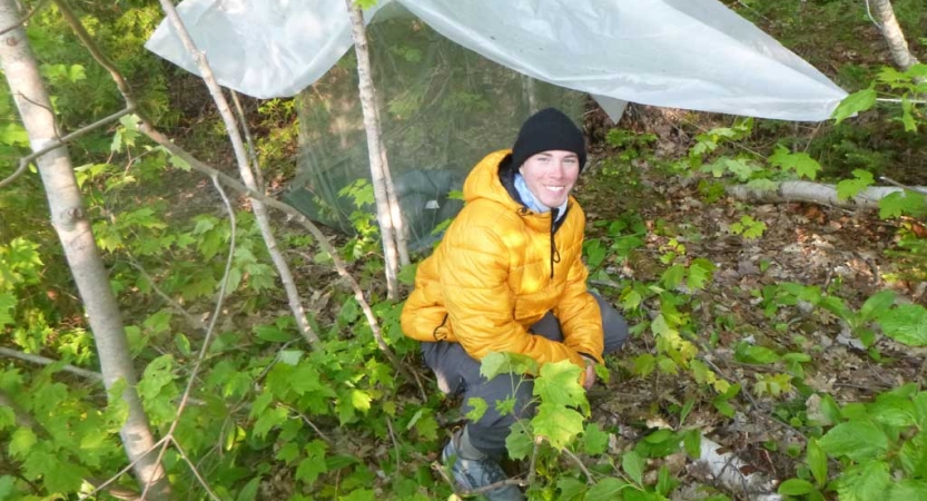 A person kneels beside a tarp shelter in a wooded area and smiles for the photo. 